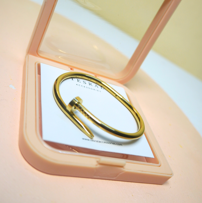 A gold-toned metal cuff ring with a flat front band, displayed inside an open peach-colored square compact on a white card. The brand name 'TESRAL' is visible on the card.