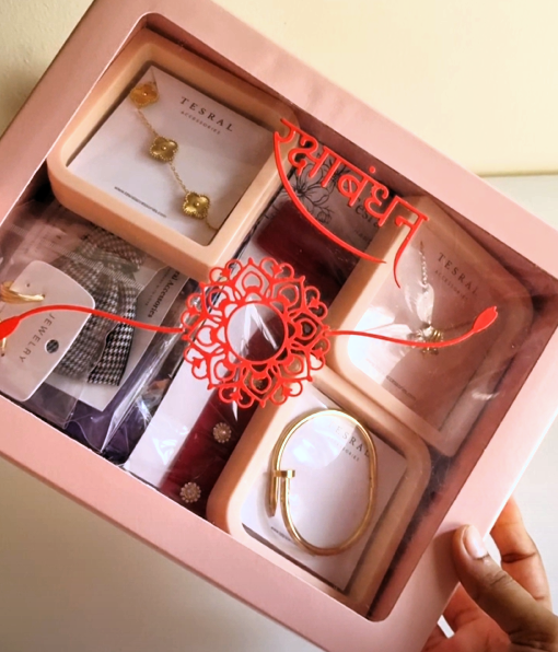 A pink gift box containing multiple jewelry items from the brand TERSAL, including gold necklaces, bracelets, and a patterned accessory, with a red decorative thread and a hand holding the box. Raksha Bandhan Gift for Sisters