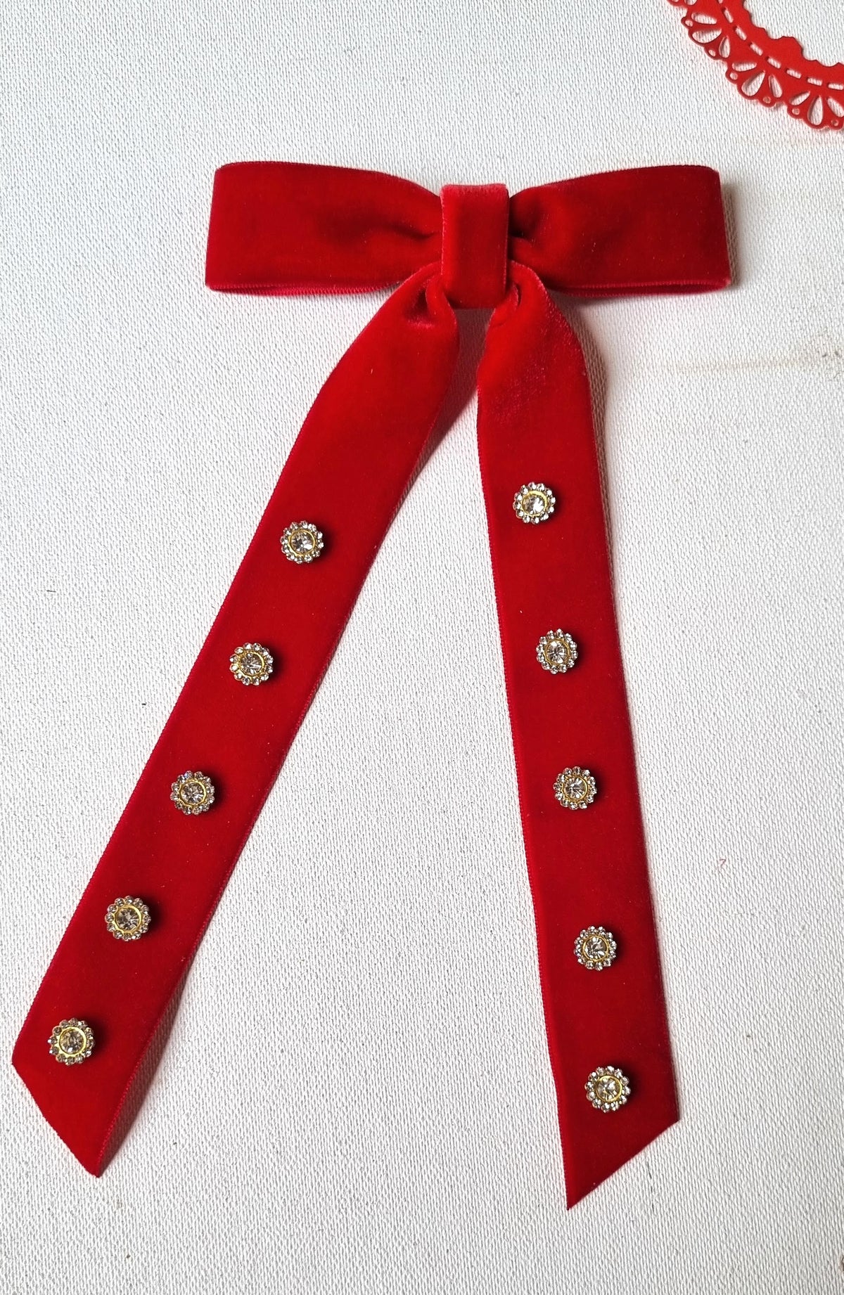 A red velvet ribbon bow with two long tails, decorated with small round silver-tone rhinestone studs, laid flat on a white textured fabric background.