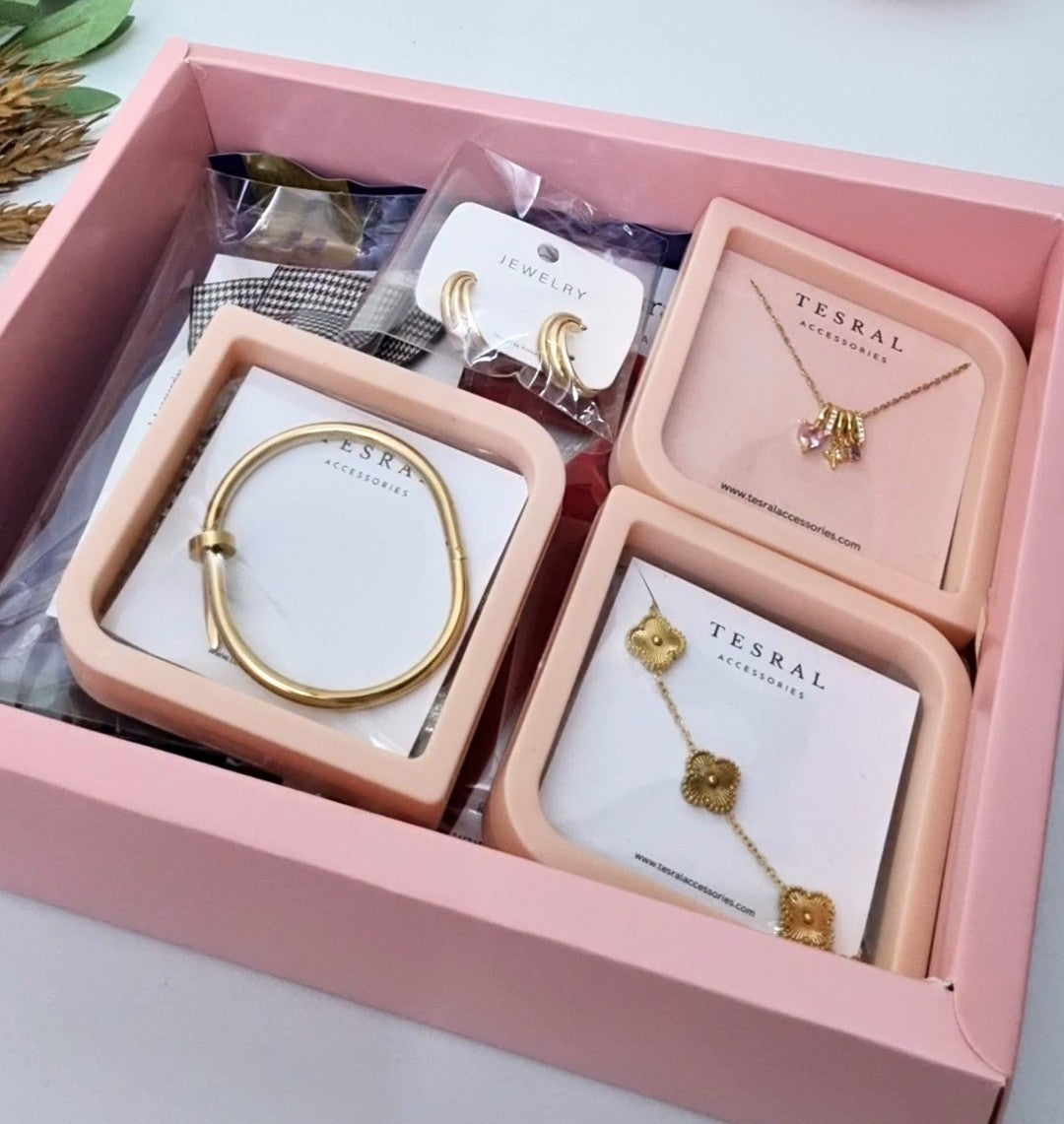 A pink jewelry gift box containing multiple gold-toned accessories from TESRAL ACCESSORIES, including a bracelet, earrings, necklace, and charm set, arranged on white cards inside a pink tray.