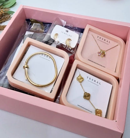 A pink jewelry gift box containing multiple gold-toned accessories from TESRAL ACCESSORIES, including a bracelet, earrings, necklace, and charm set, arranged on white cards inside a pink tray.