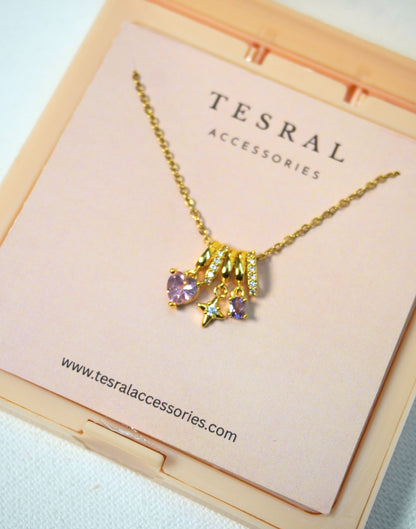 A gold-toned necklace with a cluster pendant featuring a heart-shaped purple gemstone and small clear stones, displayed on a pink card inside a peach-colored box. The card is branded TESRAL ACCESSORIES.